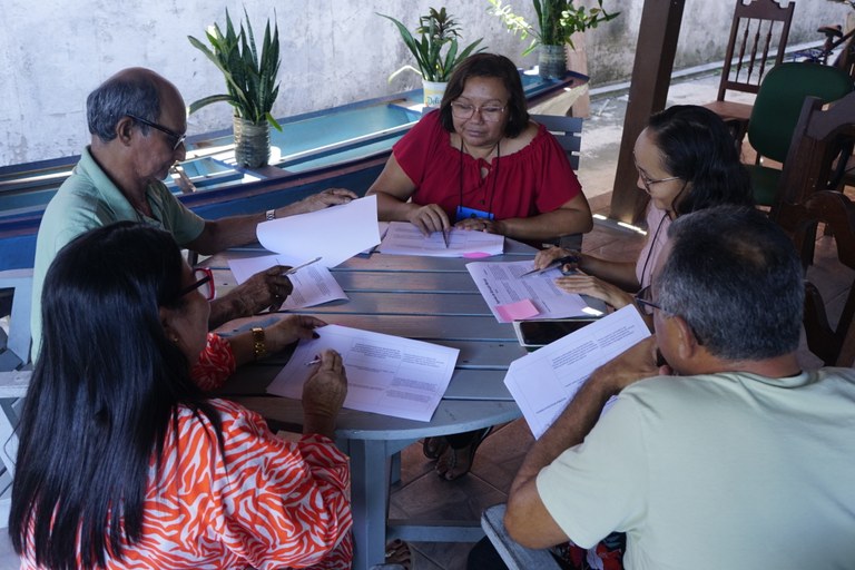 Na foto aparecem mulheres e homens sentados a uma mesa redonda analisando documentos 