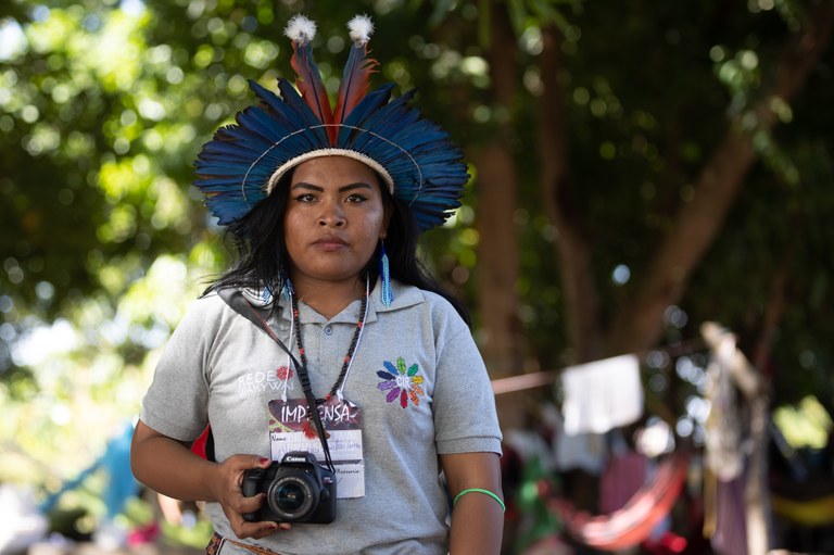 Indigenous communicator Alane dos Santos Lima, from the Macuxi people, shows a camera. She is young, wears a gray T-shirt and a headdress.