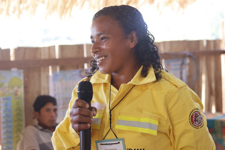 A brigadista Samara é uma indígena que está sorrindo, vestida de uniforme amarelo e tem cabelos curtos. Ela segura um microfone