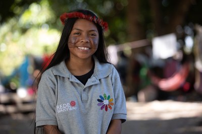 A indígena Heslen Alves da Silva sorri para a câmera. Ela tem os cabelos compridos, está de camiseta cinza e usa um cocar vermelho na cabeça