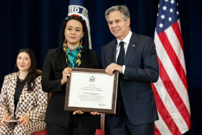 In the photo, Kari wears a dark outfit and wears a green feather ornament on her head. He holds the certificate next to Secretary Antony Blinken, who is a white, gray-haired man in a suit.