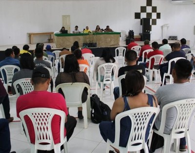 the photo shows people with their backs sitting on white chairs listening to lectures by other people at a table in the back of the room