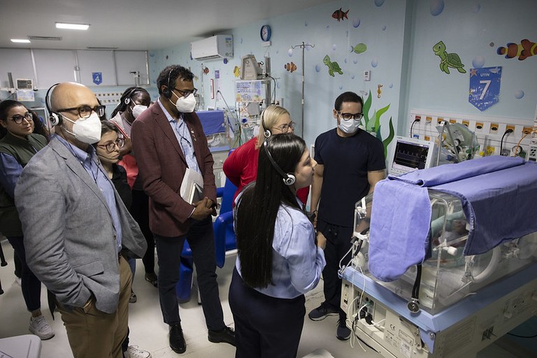 Three men using health masks looking at a baby incubator in the maternity ward that is covered with a light cloth. They are accompanied by women